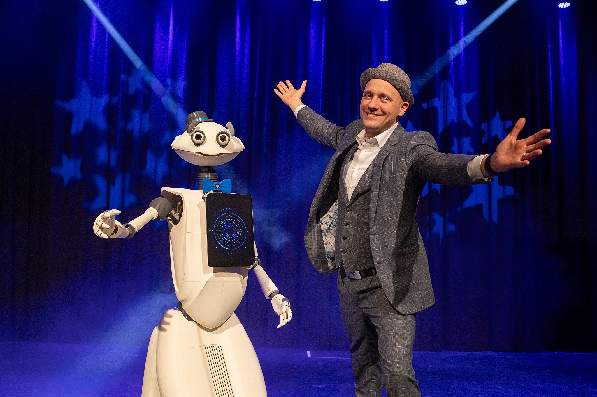 Ein Mann im grauen Anzug steht mit geöffneten Armen neben dem weißen Eventroboter Sparky, der eine blaue Fliege trägt, auf einer blau beleuchteten Bühne.